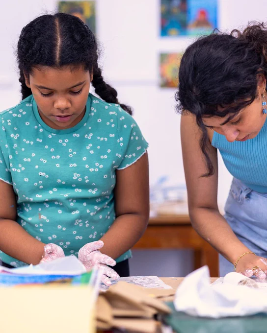 A young girl moulds something with white clay. A woman (the workshop facilitator) does the same to her right. 