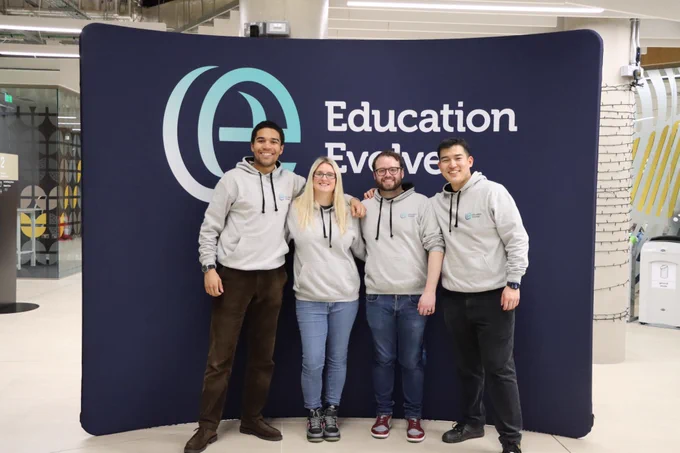 Four people wearing grey hoodies stand in front of an Education Evolved banner