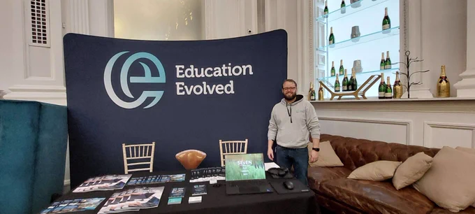 Dr Matthew Leeper stands next to a table covered in booklets and an open laptop, and in front of a banner with the Education Evolved Logo