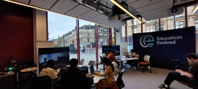 Groups of people seated around tables with laptops and large screens in a room featuring an Education Evolved banner