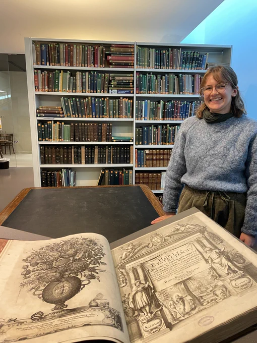 Librarian standing next to the large open book - over 50cm in size in the Garden's library, with shelves of books in the background