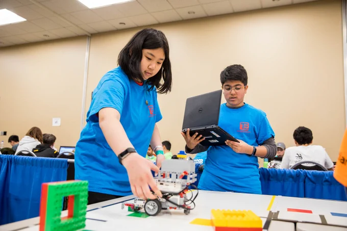 Two young people in electric blue t-shirts work together to operate a LEGO robot using a laptop.
