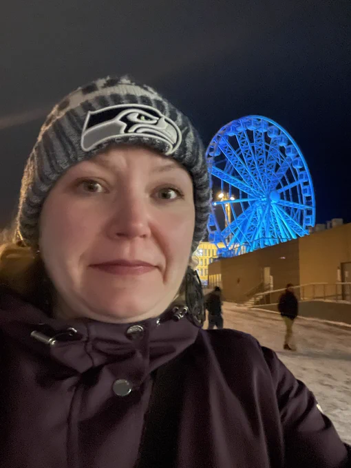 Me looking uncomfortable in a winter jacket and a Seahawks toque in front of a ferris wheel that had been lighted with blue lights