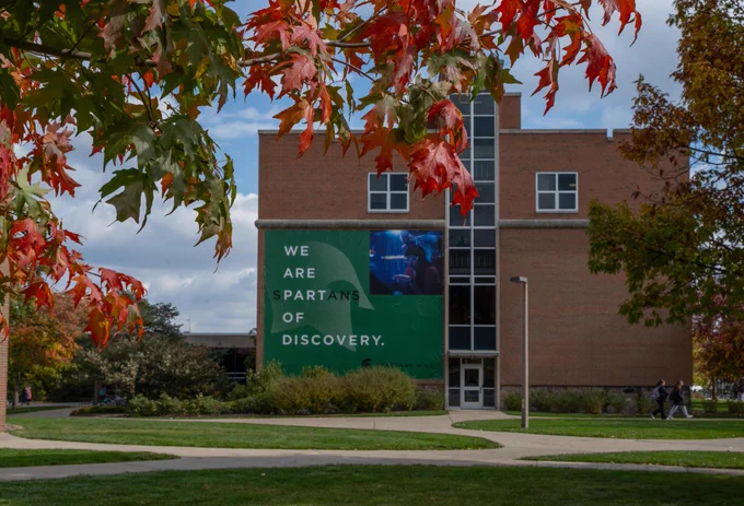 Green banner that says We are Spartans of Discovery hanging on the side of the International Student Center, which is framed by orange and red leaves