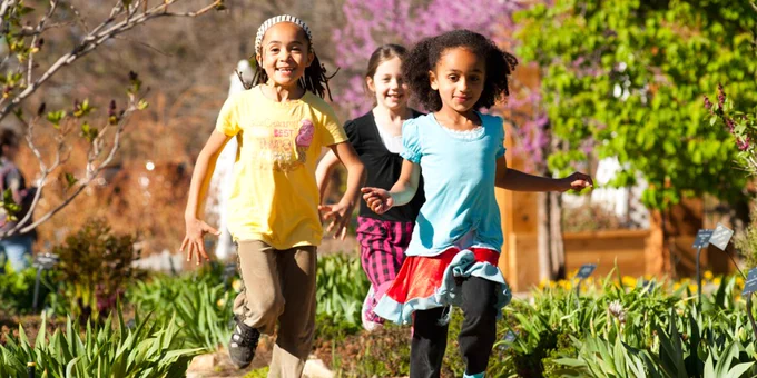 Three young children run toward the camera on a gravel path surrounded by green plants. They wear warm weather clothing of short-sleeved shirts and pants. 