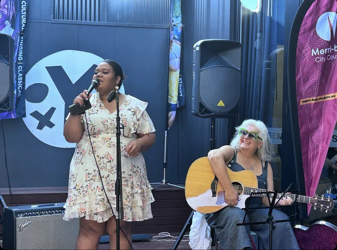  Ruci standing with microphone singing while Irine is seated, smiling and playing guitar.