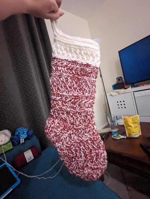 A crocheted Christmas stocking, a variety of stitches are used to create the texture. Puff stitch, alpine stitch and back loop only stitches. The majority of the stocking is made with red and white yarn together giving a mottled candy cane effect. The top is white. A plushie frog can be seen in the background.