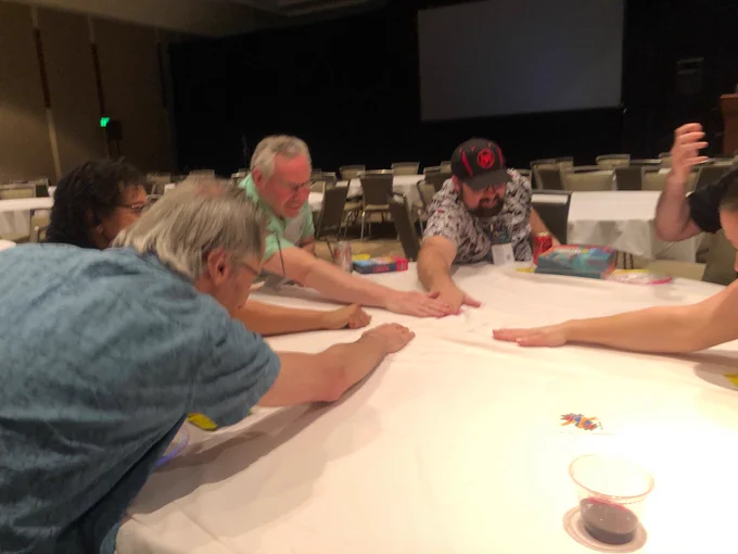A strange action photo of several people sitting at a large round table, cans of stretched toward the center