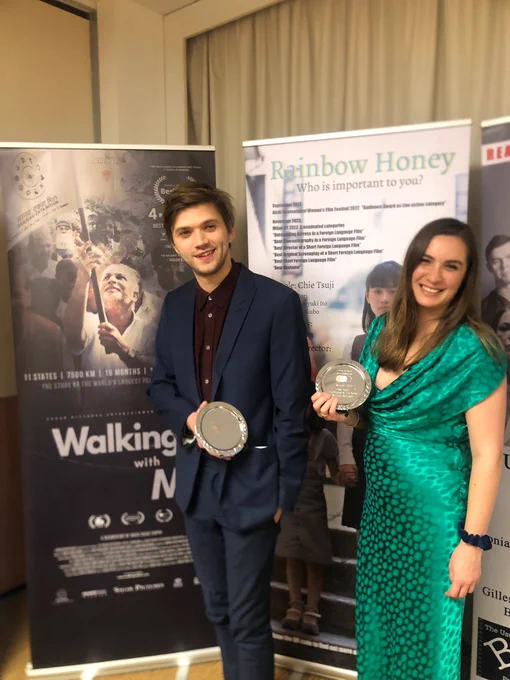 Tom, white man, 30, brown hair in a blue suit with a burgundy shirt, stands next to Ruth, 30s, shiny green dress. They're both holding plates engraved with film prizes and smiling. Behind them are two movie posters.
