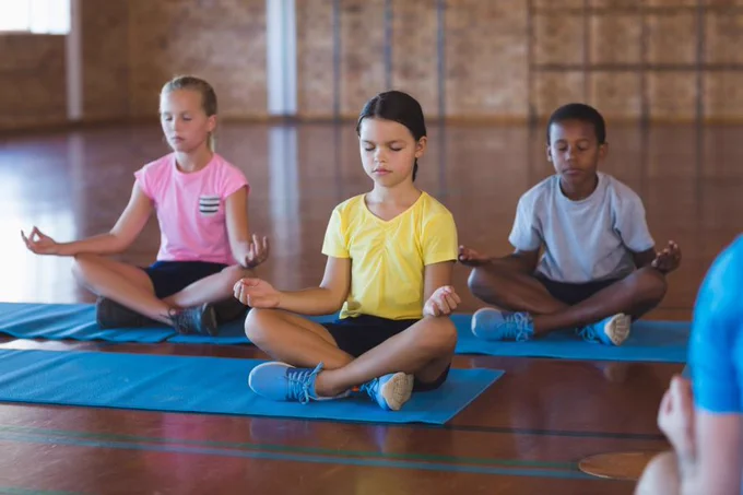 Two girls and a boy sit cross-legged, with eyes closed and hands palm up on knees, on yoga mats in a school gym. The girl in the foreground has brown hair and is wearing a yellow shirt. The girl on the left back is white with blonde hair wearing a pink shirt. The boy on the back right is Black with short hair, wearing a grey shirt.
6
