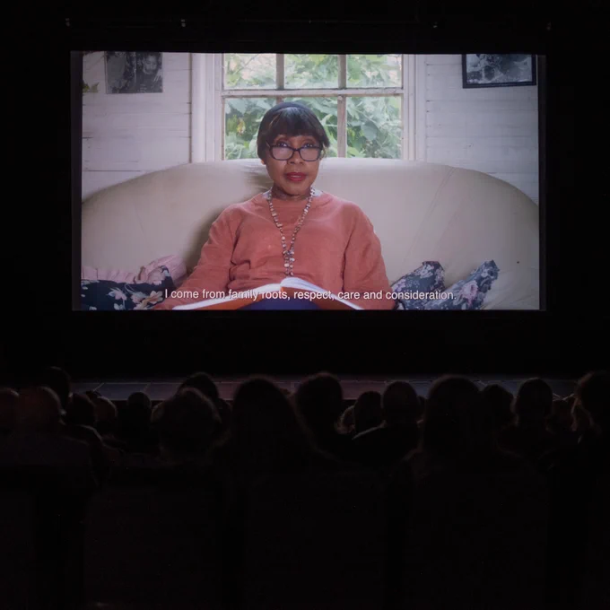 In a dark cinema, a large audience watches a projected film. On the screen, a person speaks directly to the camera. The subtitle reads: "I come from family roots, respect, care and consideration."