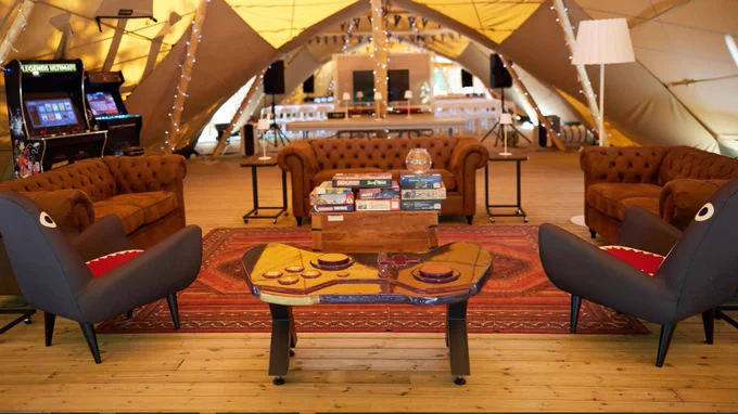 The inside of The Tent at Rare. Two shark chairs are placed either side of a table shaped like an Xbox controller. Board games and arcade cabinets can be seen in the background.
