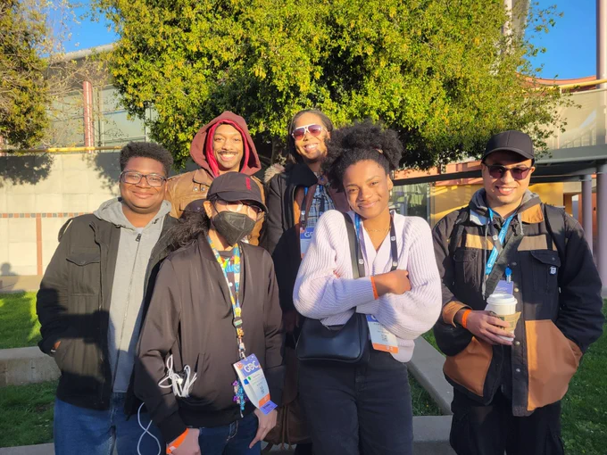 6 GADA scholars and mentors standing in front of grass, trees, and buildings outside the Game Developers Conference