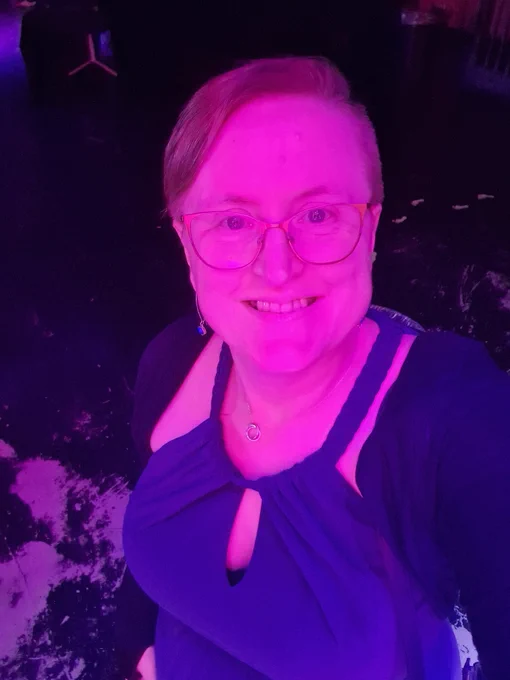 Photo of Ruth in a halter neck dress and bolero jacket smiling at the camera, bathed in pink light