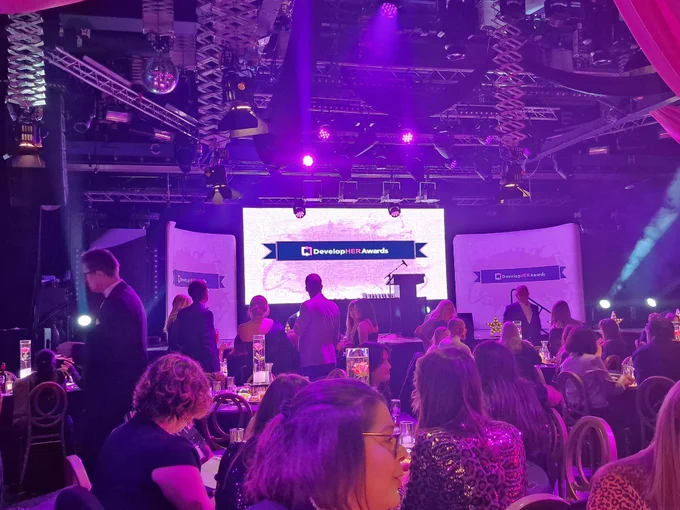 Photo of a large auditorium with the DevelopHer Awards logo showing on the main screen and two large displays on either side. The auditorium is full with circular tables and people sitting and standing chatting, illuminated with pink and purple lights