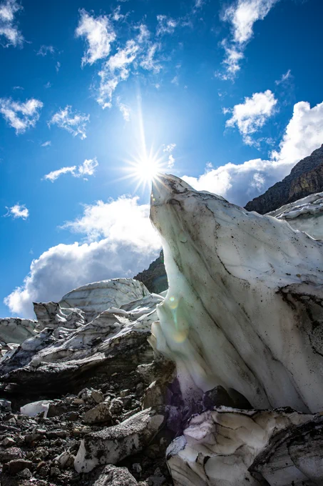 A sharp jumble of ice rises below a blue sky with some clouds and the sun.