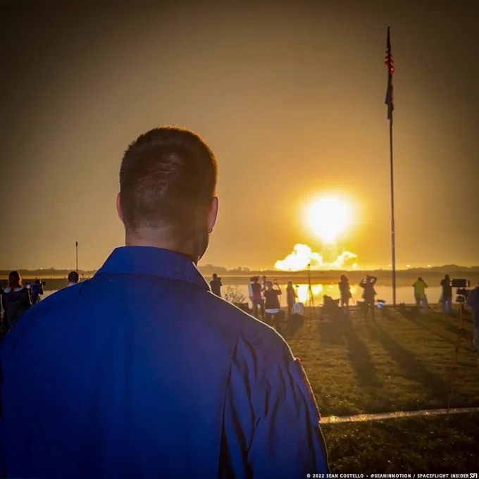 A man in a blue flight suit looks at a rocket night launch, a ball of light in the distance.