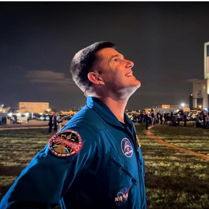 A man wearing a blue flight suit smiles as he looks up at the sky. A big building behind him sports the NASA logo.