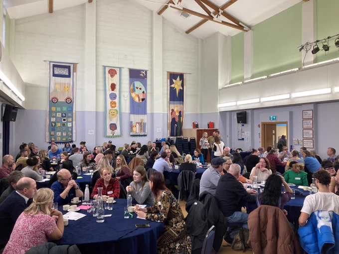 Photo from regional learning event with Changing Futures partnerships. Participants sit around 9 large tables and talk to each other.