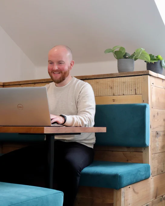A person smiling whilst sitting in a booth on a laptop. 