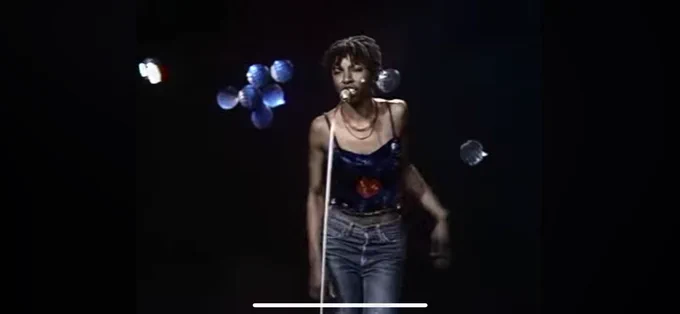 An image from a community broadcast. A woman sings into a microphone, lit under a spotlight with blue lights on a black background.