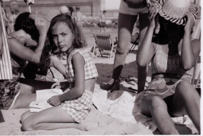 A black and white photograph of a girl on a beach looking at the camera. There are other people in the image.