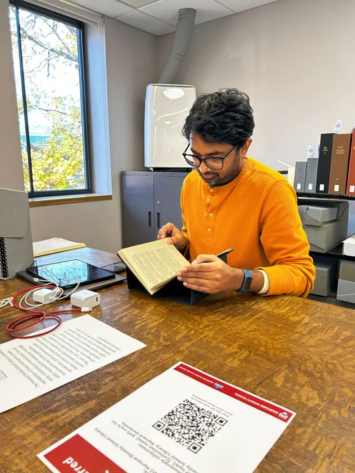 A library patron is viewing a pamphlet in a set of cradles to support the archival material. The patron is wearing glasses and is reading intently. 