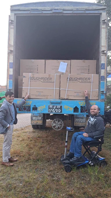 2 men in front of the back of a truck. The truck is open and filled with boxes of wheelchairs. 