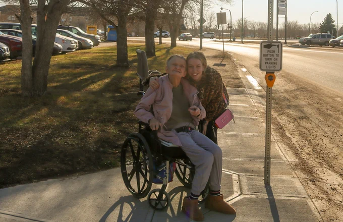 Two women, one using a wheelchair, smile at the camera beside a crosswalk button.