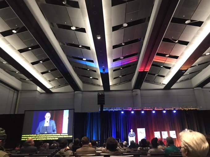 In a large conference hall, audience members in chairs on the foreground, Dr Colin Scott on the stage at a podium, with a large captioned screen to the left and a colorfully lit ceiling above.
