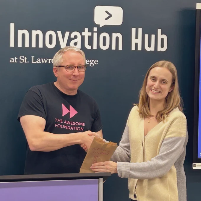 Dean of Awesome Matt Dubblestein hands over the brown paper bag of $1,000 cash to Sophia from Bayridge Secondary School for their upcoming production of High School Musical! They stand in front of the St. Lawrence College Innovation Hub logo, where we hosted the pitch party.