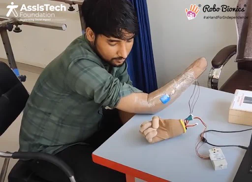 A man is sitting at a desk and is trying out a new prosthetic hand. At the top are the logos for AssisTech Foundation and Robo Bionics.