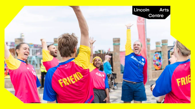 7 dancers wearing bright sports wear with the word Friend on the back are stood in a circle celebrating together. 