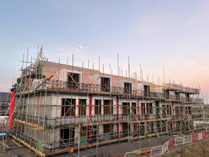 A row of houses that are being built on a construction site - scaffolding on the front, but the homes look nearly complete. Blue sky and morning sun in background.