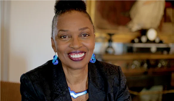 An African American woman is smiling towards the camera, wearing a black blazer and bright blue dangling earrings and a necklace. 