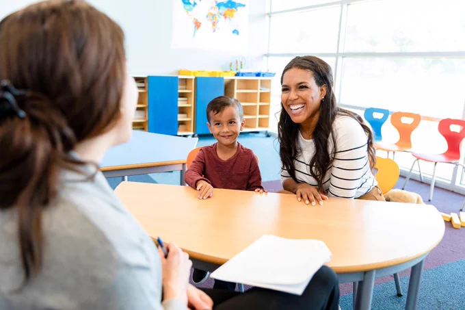 Preschool age boy in a red shirt smiles as his mother laughs across the table from a teacher (the teacher's back is facing the camera and they have brown hair in a pony tail). They are seated at a table in a classroom.