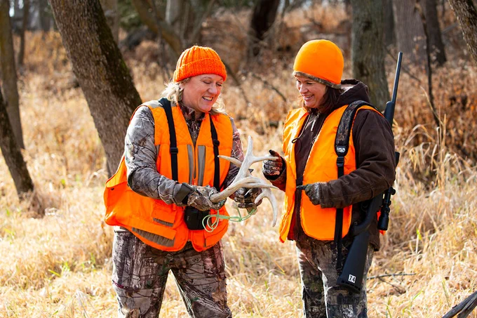 Two hunters wearing blaze orange, looking at a set of antlers while standing in the woods.