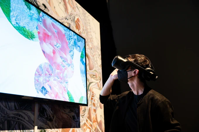 A person in a VR headset stands in front of a screen that shows a view of the virtual art installation seen inside the headset; it feature pink, blue, and green spherical forms in a white void

A visitor to the Gray Area Festival interacts with Nat Decker’s Touch, 2023. Photo: Barak Shrama