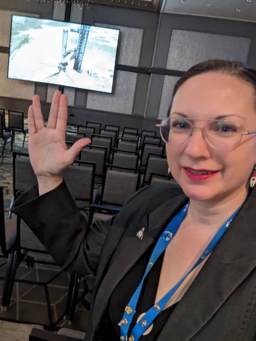 Woman wearing rocket earrings holding her hand up in the Live Long and Prosper sign that is pointing at a big screen in a mostly empty conference room.