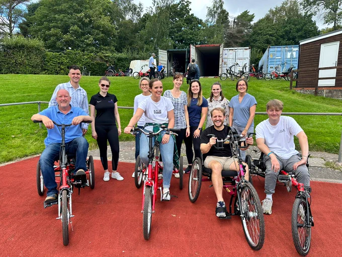Eleven of the Sense Active team posing for a photo on a running track with big smiles. Some are sat on varying forms of inclusive cycling equipment, and some are standing. 