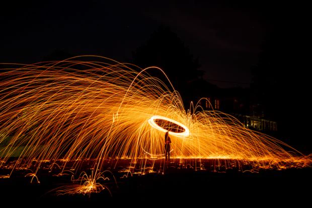 A person standing in a dark space spins burning steel wool over their head, sending a shower of orange sparks all around. 