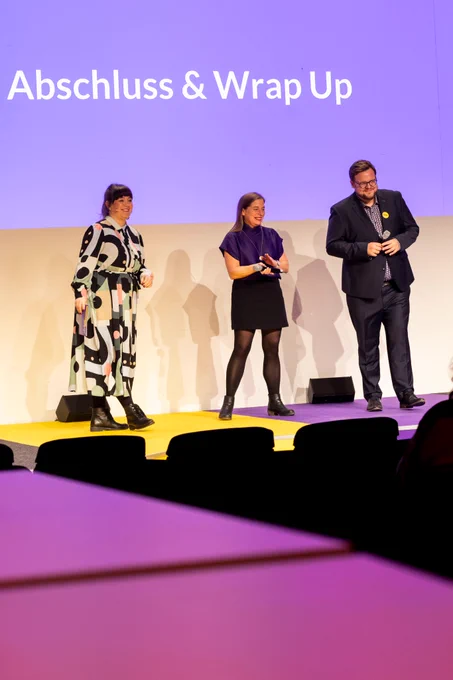 zwei frauen und ein mann stehen auf einer bühne. auf der leinwand steht abschluss und wrap up