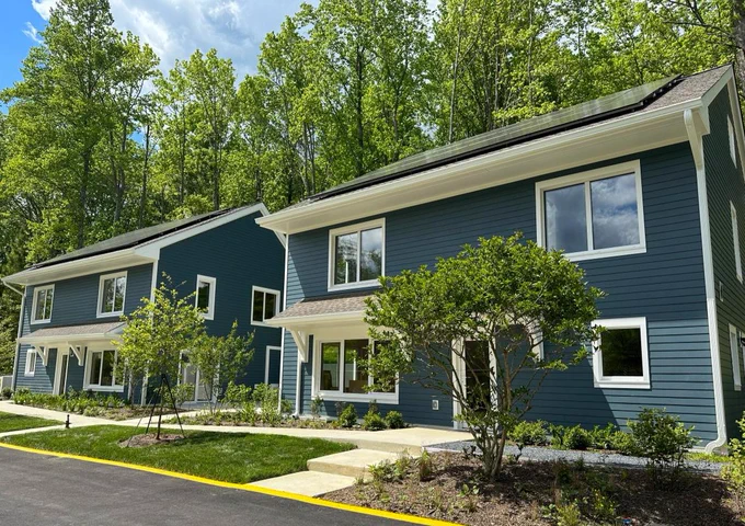 Two blue cottages with rooftop solar panels, between a road and a forest