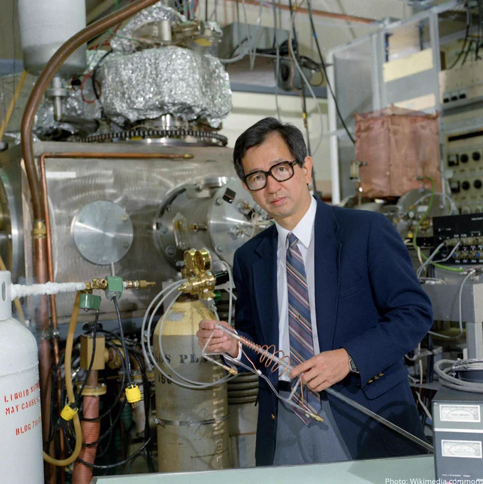 Scientist Yuan T. Lee in his lab wearing a blue blazer and a blue striped tie and grey pants. He is wearing glasses and holding a device in his hands. 