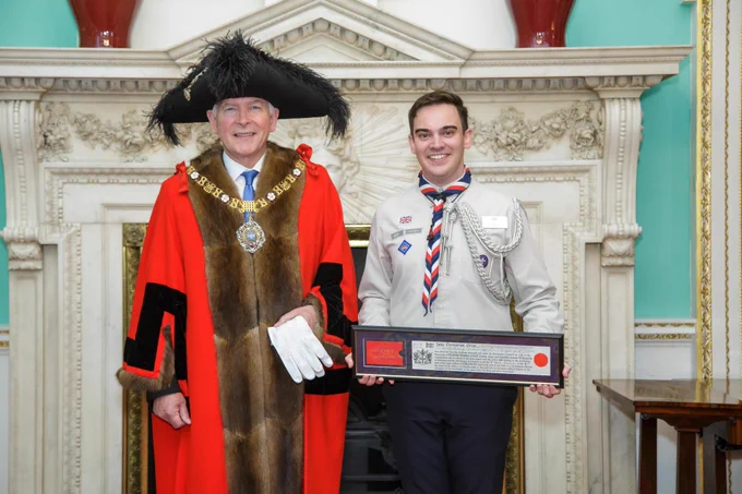 Lord Mayor Nicholas Lyons stood in scarlet Alderman's gown on the left alongside Jake in Scout uniform on the right holding his freedom certificate, in front of a fire place in the Mansion House. 