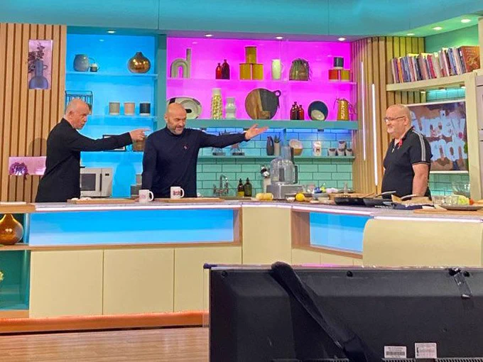 The scene is the kitchen set in Sunday Brunch.  Behind the counter are hosts Tim Lovejoy, dressed in a dark jacket with short hair, Simon Rimmer, in a dark jumper with short hair, both pointing at me, in a black polo shirt and short (no) hair! Behind are an array of items and books on shelves