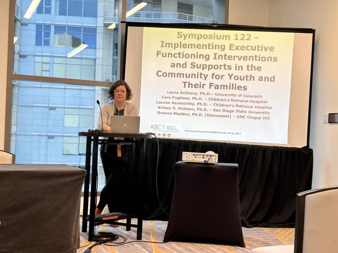 Dr. Laura Anthony presenting at a podium in front of projector screen.