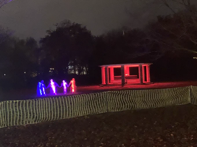 An illuminated pavilion with a group of models of people, all illuminated in different colours, set in parkland in the relative darkness, with dark trees in the background and a fence in the foreground.