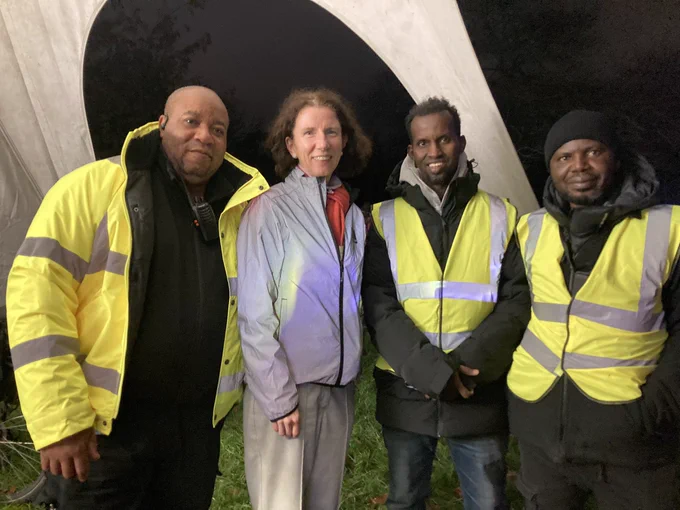 Anneliese wearing a reflective jacket accompanied with three members of Oxford Community Action all wearing reflective tabards, outside in relative darkness under part of a gazebo set up on grass.