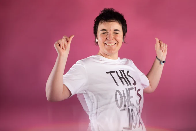 Photo of Katherina Radeva dancing in front of a pink back drop, wearing a white t-shirt with black text reading 'This One's Joy' on it.